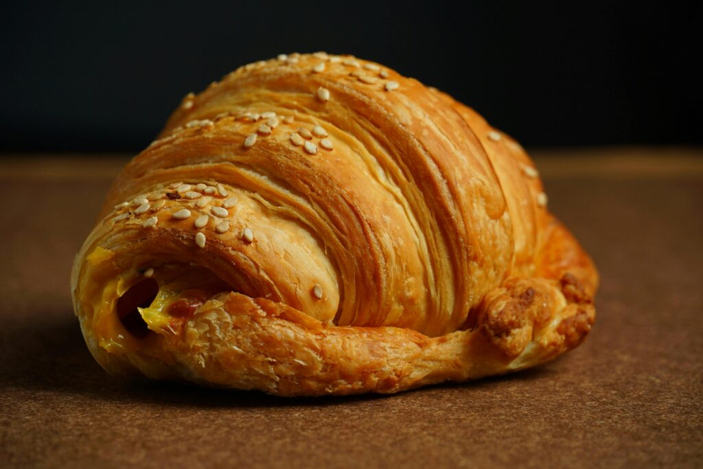 Golden brown cheese croissant sprinkled with sesame seeds on a textured surface.